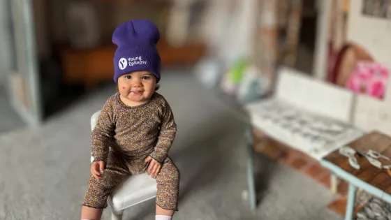 Baby girl sitting up wearing a purple Young Epilepsy beanie hat