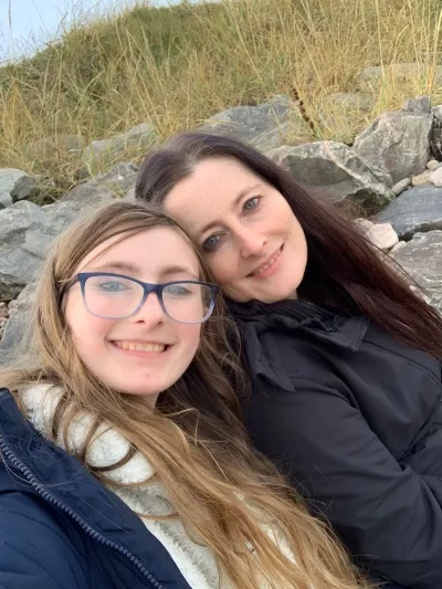 A mother and daughter in winter clothes smile in a selfie outdoors.