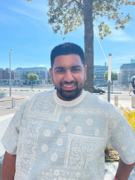 A bearded young man in a white t-shirt smiles into the camera on a summery day.