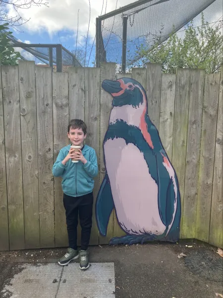A young boy, Tate, stands beside a fence with a painting of a penguin.